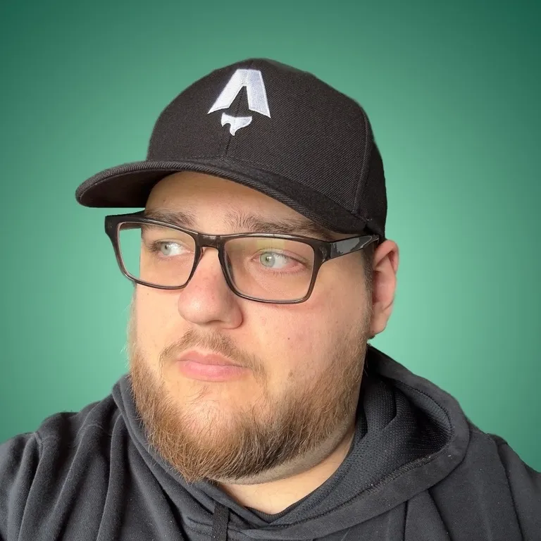A picture of Ryan, a bearded person wearing glasses and a black cap with an Astro logo, dressed in a dark hoodie, standing against a solid light green background. They are looking slightly to the side with a neutral expression.